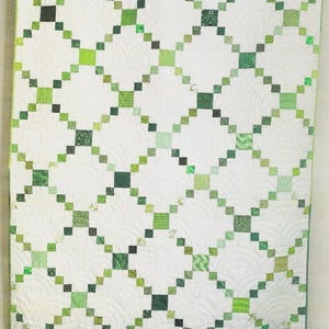 May include: A rectangular quilt featuring a white background with a repeating diagonal grid pattern composed of small squares in various shades of green. The white sections of the quilt display intricate, curved, fan-like machine quilting stitches.