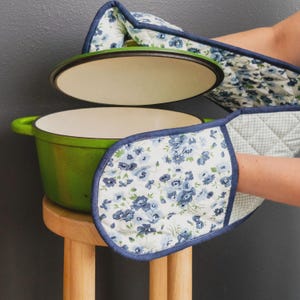 Könnte beinhalten: Ein grüner gusseiserner Schmortopf mit cremefarbener Innenseite und passendem Deckel, kombiniert mit einem gesteppten Ofenhandschuh aus weißem Stoff mit blauem Blumenmuster und dunkelblauem Saum an den Rändern des Ofenhandschuhs.