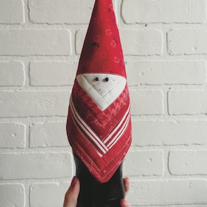 May include: A fabric gnome doll in red and white, with a button detail. The gnome features a red hat with a floral design, a white face, and a red and white striped beard. It is placed on a black bottle.