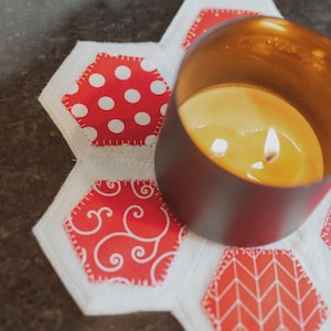 May include: A lit candle in a dark brown metal container sits on a white and red patterned hexagonal coaster. The coaster features various red patterns, including polka dots and swirls, stitched onto a white background.
