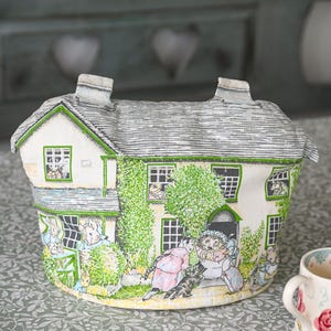 Puede incluir: Un caprichoso calentador de té con forma de casa, con una ilustración detallada de personajes animales en un jardín. El calentador tiene un tejado gris, adornos verdes y paredes blancas. El diseño incluye ventanas y una puerta.