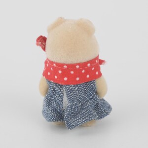 Puede incluir: Una peque&ntilde;a figura de oso beige con una bufanda roja de lunares y un vestido vaquero azul. El oso tiene un peque&ntilde;o lazo rojo en la cabeza. La figura est&aacute; sobre un fondo blanco liso.