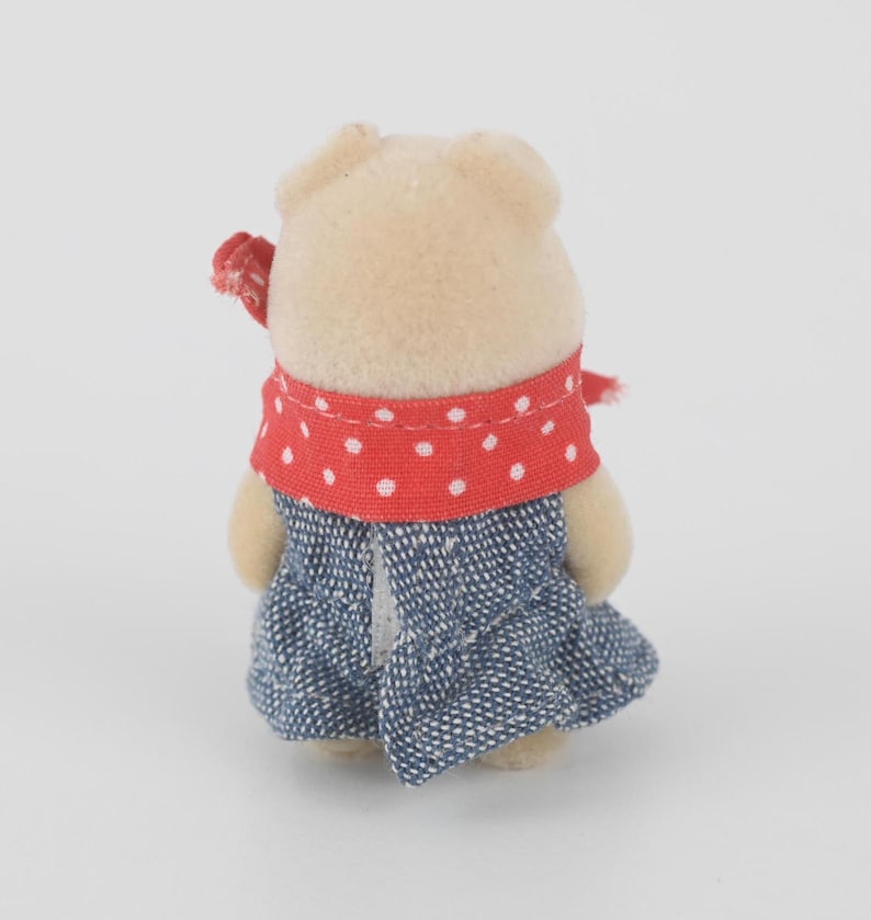 Puede incluir: Una peque&ntilde;a figura de oso beige con una bufanda roja de lunares y un vestido vaquero azul. El oso tiene un peque&ntilde;o lazo rojo en la cabeza. La figura est&aacute; sobre un fondo blanco liso.
