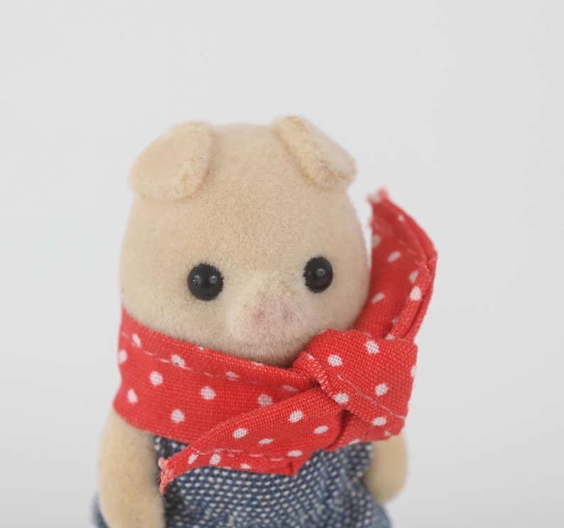 Puede incluir: Figura de cerdo beige claro con ojos negros y hocico rosa. Lleva un pa&ntilde;uelo rojo con lunares blancos y un atuendo de estilo denim azul. El juguete tiene orejas peque&ntilde;as y redondeadas.