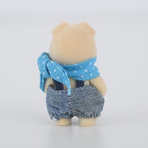 Puede incluir: Una peque&ntilde;a figura de oso de color canela, vista desde atr&aacute;s. Viste un peto vaquero con tirantes y una bufanda azul con lunares blancos. El juguete tiene orejas y pies peque&ntilde;os y redondeados.