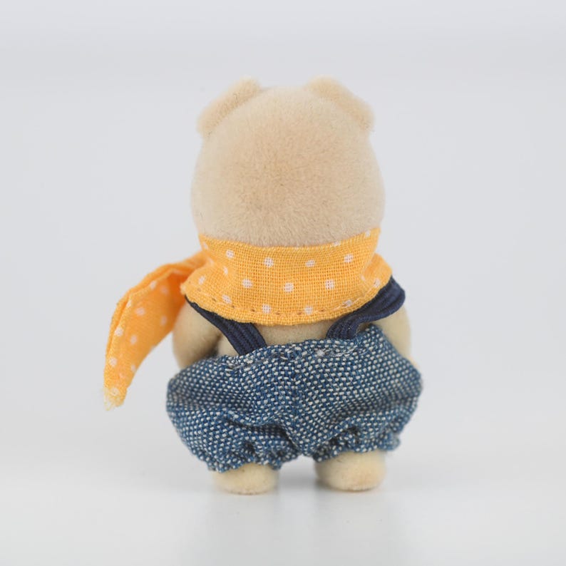 Puede incluir: Peque&ntilde;a figura de oso de peluche vista desde atr&aacute;s. El oso lleva un peto vaquero azul con tirantes azul marino y una bufanda naranja brillante con lunares blancos. El pelaje del oso es de color marr&oacute;n claro.