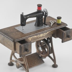 K&ouml;nnte beinhalten: Eine Miniatur-Antik-N&auml;hmaschine mit schwarzem Geh&auml;use und einem Holztisch. Der Tisch hat zwei kleine Schubladen und goldfarbene Zierelemente. Eine Spule roten Fadens sitzt auf der Maschine, mit einer gelben Spule und winzigen Scheren in der N&auml;he.