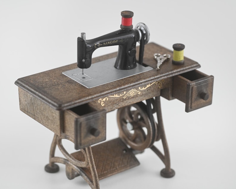 K&ouml;nnte beinhalten: Eine Miniatur-Antik-N&auml;hmaschine mit schwarzem Geh&auml;use und einem Holztisch. Der Tisch hat zwei kleine Schubladen und goldfarbene Zierelemente. Eine Spule roten Fadens sitzt auf der Maschine, mit einer gelben Spule und winzigen Scheren in der N&auml;he.
