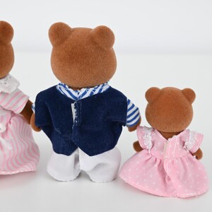 May include: Three brown bear figurines. One wears a pink and white striped dress with a white collar and bow. Another wears a navy blue vest over a white shirt. The third bear wears a pink dress with white polka dots.