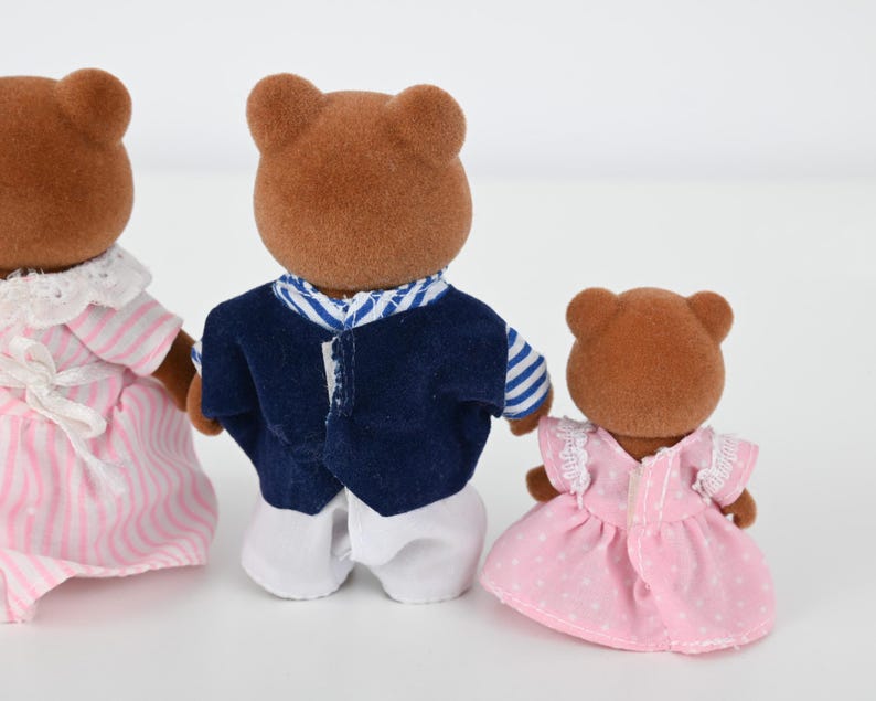May include: Three brown bear figurines. One wears a pink and white striped dress with a white collar and bow. Another wears a navy blue vest over a white shirt. The third bear wears a pink dress with white polka dots.