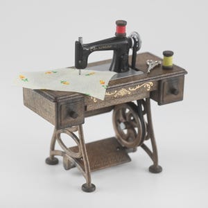 K&ouml;nnte beinhalten: Miniatur-Singer-N&auml;hmaschine mit schwarzem Geh&auml;use, roter Spule und Holztisch. Der Tisch hat zwei Schubladen und ein goldenes Dekor. Ein Stoffst&uuml;ck mit Blumenmuster liegt unter der Nadel.