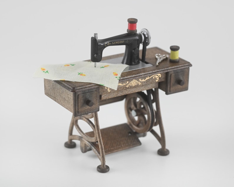 K&ouml;nnte beinhalten: Miniatur-Singer-N&auml;hmaschine mit schwarzem Geh&auml;use, roter Spule und Holztisch. Der Tisch hat zwei Schubladen und ein goldenes Dekor. Ein Stoffst&uuml;ck mit Blumenmuster liegt unter der Nadel.