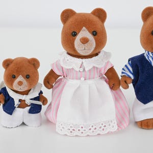 May include: Three brown bear figurines. The bear in the middle wears a pink and white striped dress with a white apron. The other two bears wear blue and white outfits. The bears have white faces and black eyes.