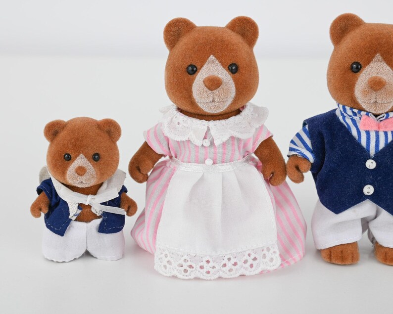 May include: Three brown bear figurines. The bear in the middle wears a pink and white striped dress with a white apron. The other two bears wear blue and white outfits. The bears have white faces and black eyes.