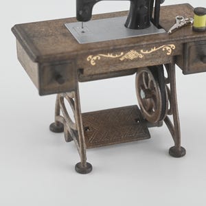 K&ouml;nnte beinhalten: Eine Miniatur-Antik-N&auml;hmaschine mit einem braunen Holztisch und schwarzen Maschinenteilen. Der Tisch hat zwei kleine Schubladen, dekorative goldene Akzente und ein Fu&szlig;pedal. Eine Schere und eine gelbe Garnrolle liegen auf dem Tisch.