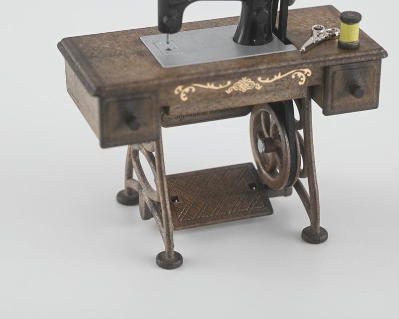 K&ouml;nnte beinhalten: Eine Miniatur-Antik-N&auml;hmaschine mit einem braunen Holztisch und schwarzen Maschinenteilen. Der Tisch hat zwei kleine Schubladen, dekorative goldene Akzente und ein Fu&szlig;pedal. Eine Schere und eine gelbe Garnrolle liegen auf dem Tisch.