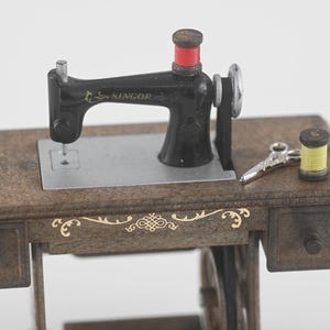 K&ouml;nnte beinhalten: Eine Miniatur-Antik-N&auml;hmaschine mit schwarzem Geh&auml;use und Holzsockel. Die Maschine hat eine rote Garnspule, eine silberne Nadel und eine kleine Schere. Der Holzsockel hat dekorative goldene Akzente und zwei kleine Schubladen.