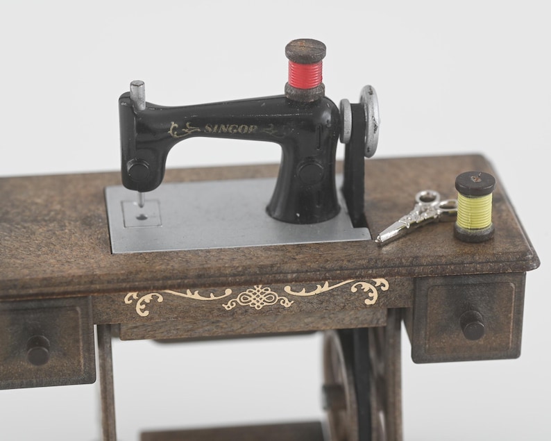 K&ouml;nnte beinhalten: Eine Miniatur-Antik-N&auml;hmaschine mit schwarzem Geh&auml;use und Holzsockel. Die Maschine hat eine rote Garnspule, eine silberne Nadel und eine kleine Schere. Der Holzsockel hat dekorative goldene Akzente und zwei kleine Schubladen.