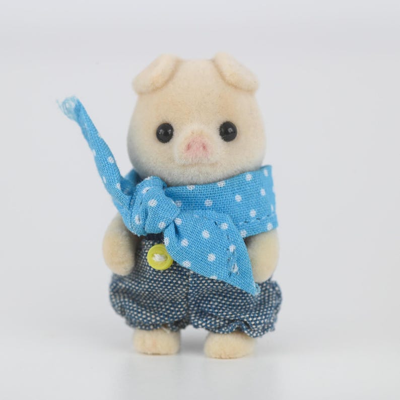 Puede incluir: Una peque&ntilde;a figura de cerdo de color claro, vestida con un peto vaquero y una bufanda azul con lunares blancos. El peto tiene un bot&oacute;n amarillo. El cerdo tiene ojos negros y una nariz rosa.