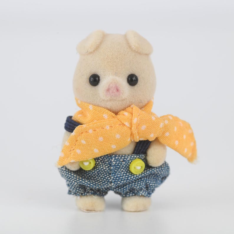 Puede incluir: Peque&ntilde;a figura de cerdo de peluche con cuerpo de color beige claro, ojos negros y nariz rosa. Viste un peto vaquero con botones amarillos y una bufanda amarilla con lunares blancos. El cerdito tiene orejas peque&ntilde;as y redondeadas.