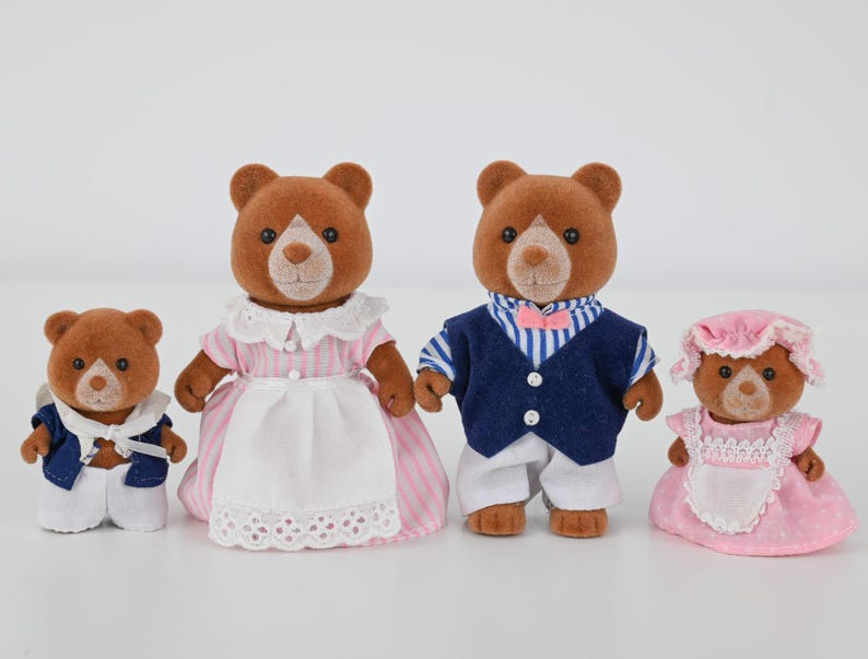 May include: Four brown bear figurines dressed in various outfits. The family includes a father in a blue vest and striped shirt, a mother in a pink dress, a child in a sailor outfit, and a baby in a pink dress and bonnet.