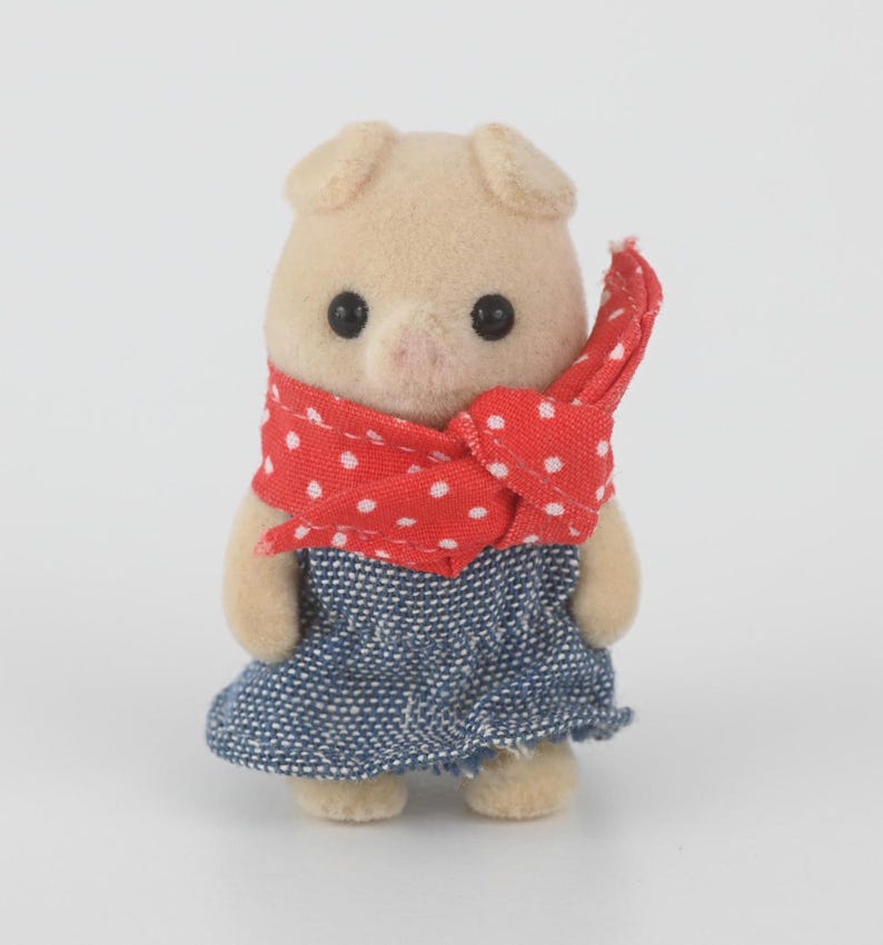 Puede incluir: Peque&ntilde;a figura de cerdo beige, con un pa&ntilde;uelo rojo de lunares blancos y un vestido vaquero azul. El cerdo tiene ojos negros y un hocico rosa. La figura est&aacute; de pie sobre un fondo blanco.