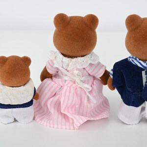 May include: Three brown bear figurines. One bear wears a white and navy outfit, another wears a pink and white striped dress, and the third wears a white and navy outfit. The bears are standing on a white surface.