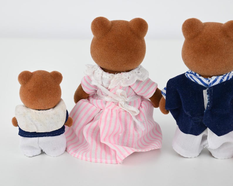 May include: Three brown bear figurines. One bear wears a white and navy outfit, another wears a pink and white striped dress, and the third wears a white and navy outfit. The bears are standing on a white surface.