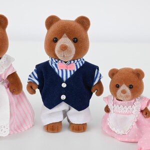 May include: Three brown bear figurines. One bear wears a navy blue vest, white pants, and a striped shirt with a pink bow tie. Another bear wears a pink and white striped dress. The third bear wears a pink dress with a white apron.