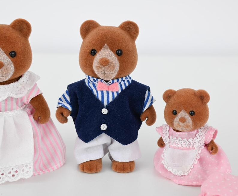 May include: Three brown bear figurines. One bear wears a navy blue vest, white pants, and a striped shirt with a pink bow tie. Another bear wears a pink and white striped dress. The third bear wears a pink dress with a white apron.