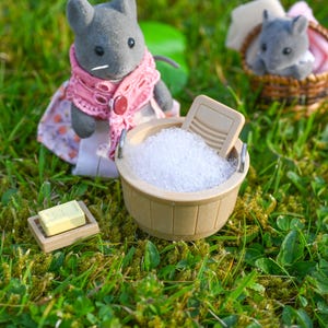 May include: A miniature scene featuring a gray rabbit figure wearing a pink shawl and dress, with a wooden washtub filled with suds. A bar of soap rests on a small tray, and another rabbit figure sits in a basket. The scene is set on green grass.