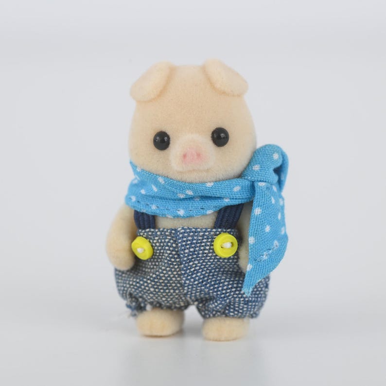 Puede incluir: Una peque&ntilde;a figura de cerdo de color beige claro que lleva un peto vaquero con botones amarillos y una bufanda azul con lunares blancos. El cerdo tiene ojos negros y una nariz rosa.