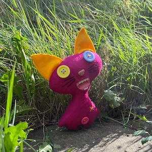 May include: A pink felt cat toy with yellow ears and a blue and yellow button eyes. The cat has a wide open mouth and a pink tongue.