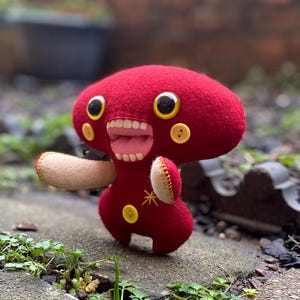 Puede incluir: Un juguete de peluche hecho a mano con forma de hongo rojo. Presenta ojos grandes y expresivos, una boca abierta con dientes y detalles de botones. El juguete tiene un diseño caprichoso con un brazo cosido y una estrella.