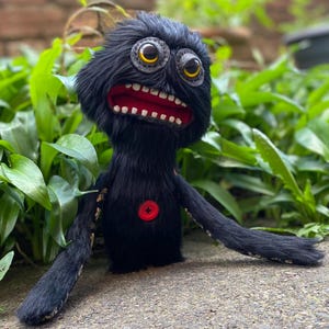 Könnte beinhalten: Ein handgefertigtes schwarzes Plüschmonster mit großen, ausdrucksstarken Augen und einem breiten, offenen Mund, der weiße Zähne und ein rotes Inneres zeigt. Es hat lange, schlanke Arme und einen roten Knopf auf der Brust. Die Kreatur steht vor einem Hintergrund aus grünem Laub.