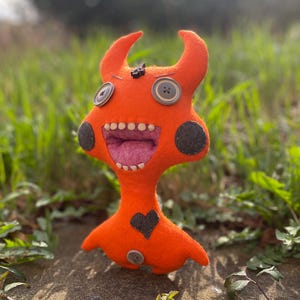Peut inclure: Jouet monstre en feutre orange avec des yeux noirs, un grand sourire aux dents et un cœur noir sur la poitrine. Le monstre a des cornes et se tient sur une surface herbeuse.