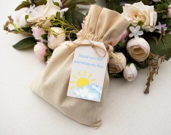 Könnte beinhalten: Eine kleine, beige Stofftasche mit einem weißen Etikett. Das Etikett zeigt eine gelbe Sonne mit blauen Wolken und den Text "Danke, dass du meinen Tag erhellt hast!"