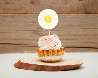 May include: A white cupcake with pink and white frosting and colorful sprinkles topped with a yellow sun with a smiley face on a wooden slice.