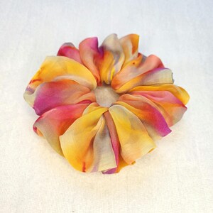 May include: A colorful scrunchie made of fabric with a floral design. The scrunchie features a blend of yellow, orange, pink, and purple hues. The fabric is gathered and folded to create a flower-like shape, perfect for accessorizing hair.