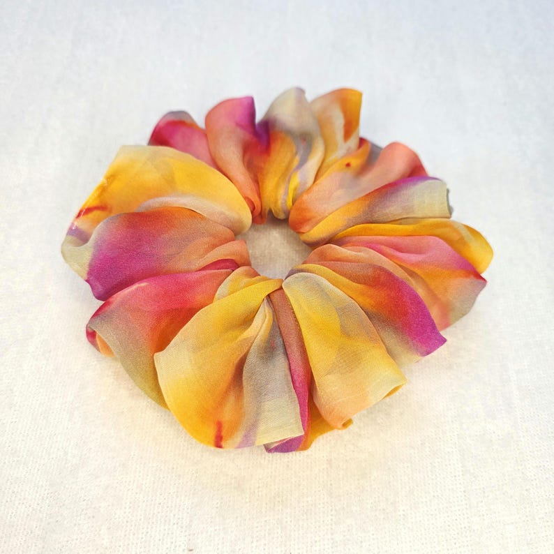 May include: A colorful scrunchie made of fabric with a floral design. The scrunchie features a blend of yellow, orange, pink, and purple hues. The fabric is gathered and folded to create a flower-like shape, perfect for accessorizing hair.