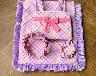 May include: A matching set of accessories in a gingham pattern of pink, purple, and orange. The set includes a tote bag with handles, a small pouch with a pink bow, a headband, and a scrunchie. The edges of the tote bag are trimmed with a ruffled purple satin.