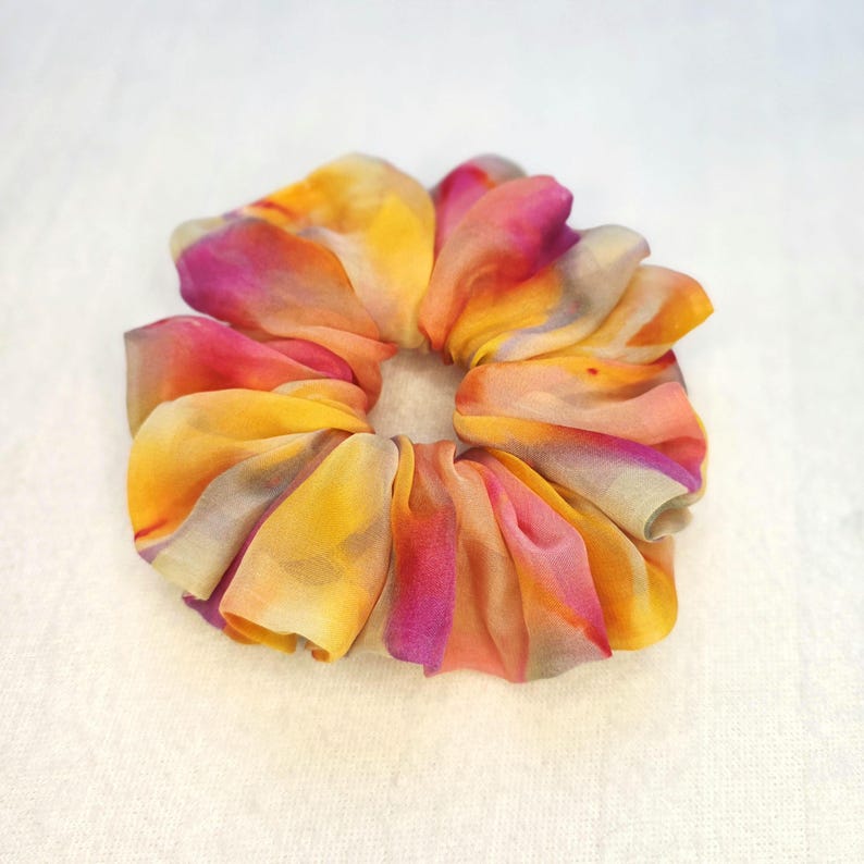 May include: A colorful scrunchie with a vibrant watercolor design. The fabric features a blend of yellow, orange, pink, and purple hues. The scrunchie is made of soft, flowing material, perfect for accessorizing hairstyles.
