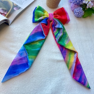 Può includere: Un fiocco per capelli colorato con motivo arcobaleno con un grande fiocco al centro e due code lunghe.