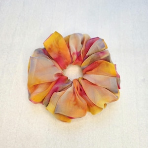 May include: A colorful scrunchie with a tie-dye effect. The fabric is a blend of yellow, orange, pink, and purple hues. The scrunchie is made of a soft, flowing material, perfect for securing hair in a ponytail or bun.