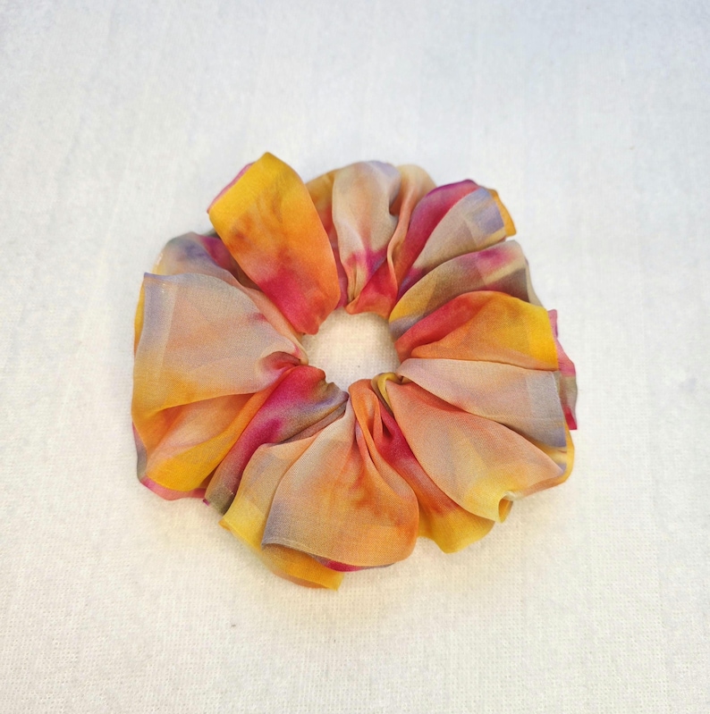 May include: A colorful scrunchie with a tie-dye effect. The fabric is a blend of yellow, orange, pink, and purple hues. The scrunchie is made of a soft, flowing material, perfect for securing hair in a ponytail or bun.