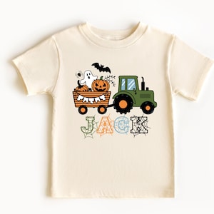 Może przedstawiać: Kremowy t-shirt z krótkim rękawem z grafiką zielonego traktora ciągnącego przyczepę wypełnioną dyniami, duszkiem, pająkiem i nietoperzem. Pod traktorem nadrukowano imię JACK dużymi literami z nałożonym wzorem pajęczyn.