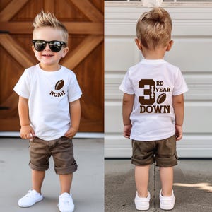 May include: A young child wearing a white t-shirt with a football graphic and the name "Noah". The child is also wearing brown shorts, white sneakers, and sunglasses. The back of the shirt says "3RD YEAR DOWN" with a football graphic.