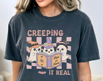 May include: Dark grey t-shirt with a Halloween graphic. The design shows three cartoon ghosts in hats, reading a book with "Trick or Teach". The shirt also has the text "Creeping it Real" on a pink and white checkered background.