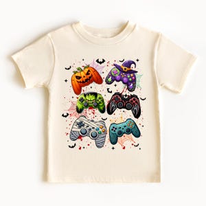 Op de afbeelding: Crèmekleurig T-shirt met zes kleurrijke Halloween-thema gamecontroller-ontwerpen. De controllers bevatten een pompoen, een heks, een Frankenstein-monster en anderen. Vleermuizen, spinnenwebben en rode spatten maken het spookachtige ontwerp compleet.
