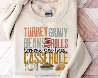 Puede incluir: Una sudadera de color arena con la frase "TURKEY GRAVY BEANS AND ROLLS lemme see that CASSEROLE" en varios colores. El diseño incluye ilustraciones de un plato, una tarta y un pavo asado.